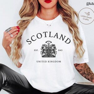 Pode incluir: Camiseta branca com a palavra "SCOTLAND" em preto, arqueada na parte superior. Abaixo, um bras&atilde;o detalhado em preto e branco e o texto "EST. 843 UNITED KINGDOM".