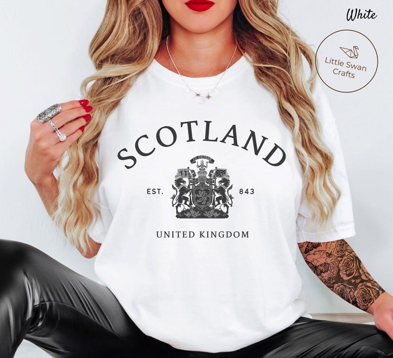 Pode incluir: Camiseta branca com a palavra "SCOTLAND" em preto, arqueada na parte superior. Abaixo, um bras&atilde;o detalhado em preto e branco e o texto "EST. 843 UNITED KINGDOM".