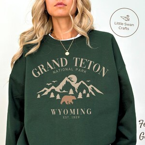May include: Forest green sweatshirt with a graphic design of a mountain range, a bear, and the text "Grand Teton National Park, Wyoming Est. 1929".