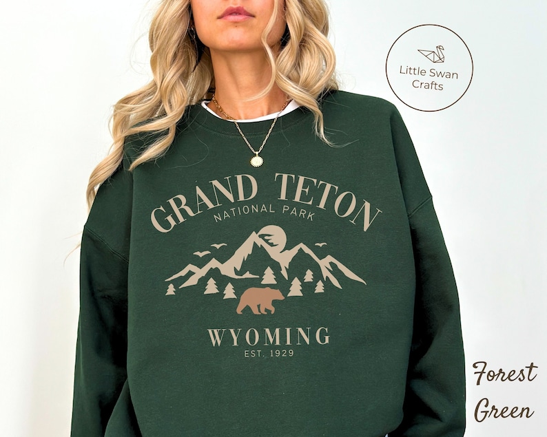 May include: Forest green sweatshirt with a graphic design of a mountain range, a bear, and the text "Grand Teton National Park, Wyoming Est. 1929".