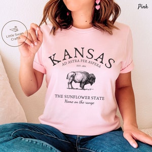 May include: Pink t-shirt with a graphic design featuring the text "KANSAS", "AD ASTRA PER ASPERA", "EST. 1861", "THE SUNFLOWER STATE", and "Home on the range". The graphic also includes a drawing of a buffalo.