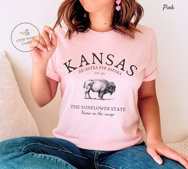 May include: Pink t-shirt with a graphic design featuring the text "KANSAS", "AD ASTRA PER ASPERA", "EST. 1861", "THE SUNFLOWER STATE", and "Home on the range". The graphic also includes a drawing of a buffalo.