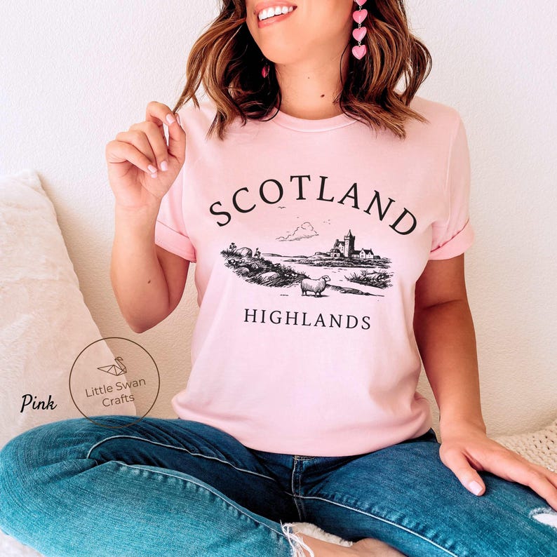 May include: Pale pink t-shirt with rolled sleeves. It features a black and white illustration of a Scottish Highlands scene, including a castle, sheep, and the words "SCOTLAND HIGHLANDS". The shirt also has the text "Pink" and a logo.