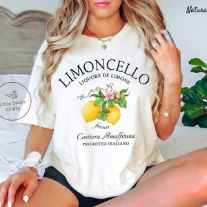 May include: White t-shirt with a graphic design of lemons and flowers. The text "Limoncello Liquore de Limone" is printed in black above the lemons. The text "Fresca Costiera Amalfitana Prodotto Italiano" is printed in black below the lemons.