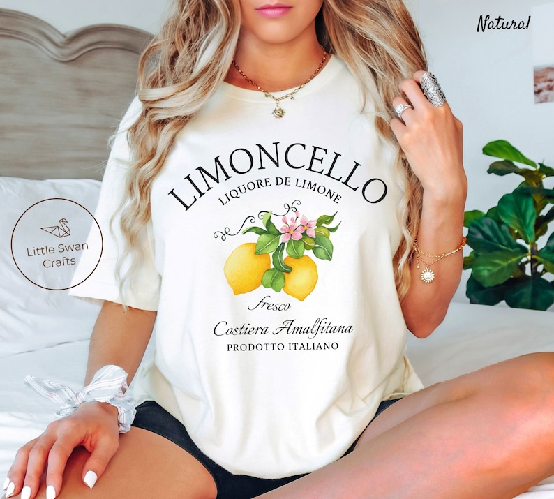 May include: White t-shirt with a graphic design of lemons and flowers. The text "Limoncello Liquore de Limone" is printed in black above the lemons. The text "Fresca Costiera Amalfitana Prodotto Italiano" is printed in black below the lemons.