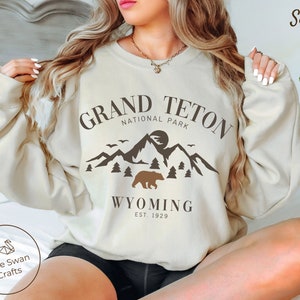 May include: A beige sweatshirt with a brown graphic design featuring a mountain range, a bear, and the text "Grand Teton National Park, Wyoming Est. 1929".
