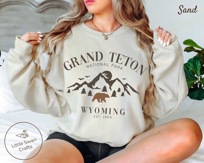 May include: A beige sweatshirt with a brown graphic design featuring a mountain range, a bear, and the text "Grand Teton National Park, Wyoming Est. 1929".