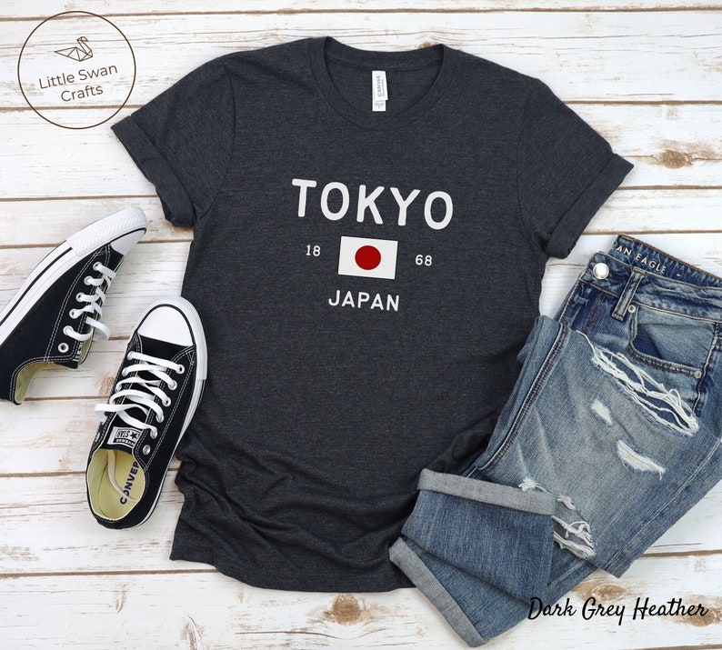 May include: Dark grey heather t-shirt with white text that reads "TOKYO" with the Japanese flag and "JAPAN" underneath. The shirt is laid out on a white wooden surface with a pair of black Converse sneakers and blue jeans.