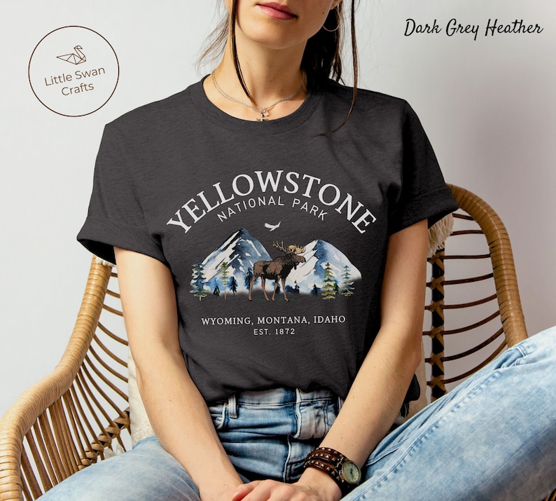 May include: A dark gray heather t-shirt with a white graphic print of Yellowstone National Park. The graphic includes a moose, mountains, trees, and the text 'Yellowstone National Park' and 'Wyoming, Montana, Idaho Est. 1872'.