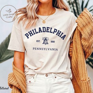 May include: A white t-shirt with a navy blue graphic of the Liberty Bell and the text "PHILADELPHIA EST. 1682 PENNSYLVANIA".