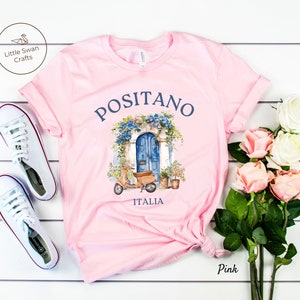 Puede incluir: Camiseta rosa con un gr&aacute;fico de una puerta azul y un scooter. El texto "Positano Italia" est&aacute; impreso en la camiseta.