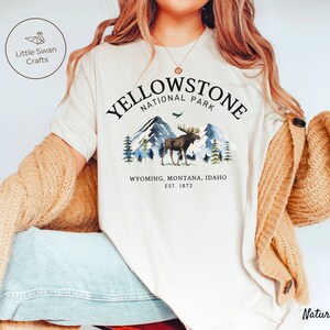 May include: Cream-colored t-shirt featuring a Yellowstone National Park design. The graphic includes a moose, mountains, and the text "Yellowstone National Park, Wyoming, Montana, Idaho, Est. 1872." The shirt is part of a clothing collection.