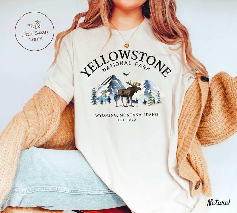 May include: Cream-colored t-shirt featuring a Yellowstone National Park design. The graphic includes a moose, mountains, and the text "Yellowstone National Park, Wyoming, Montana, Idaho, Est. 1872." The shirt is part of a clothing collection.