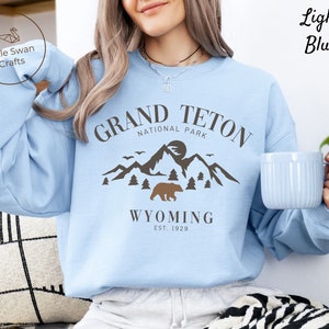 May include: Light blue sweatshirt with a graphic design featuring the text "Grand Teton National Park" and a mountain scene with a bear. The text "Wyoming Est. 1929" is below the graphic.