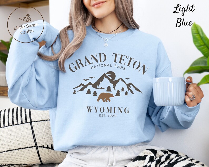 May include: Light blue sweatshirt with a graphic design featuring the text "Grand Teton National Park" and a mountain scene with a bear. The text "Wyoming Est. 1929" is below the graphic.