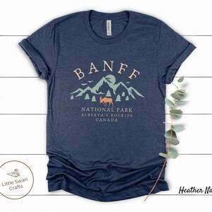 May include: Heather navy blue t-shirt with a graphic design of mountains, a moose, and the text "Banff National Park, Alberta's Rockies, Canada".