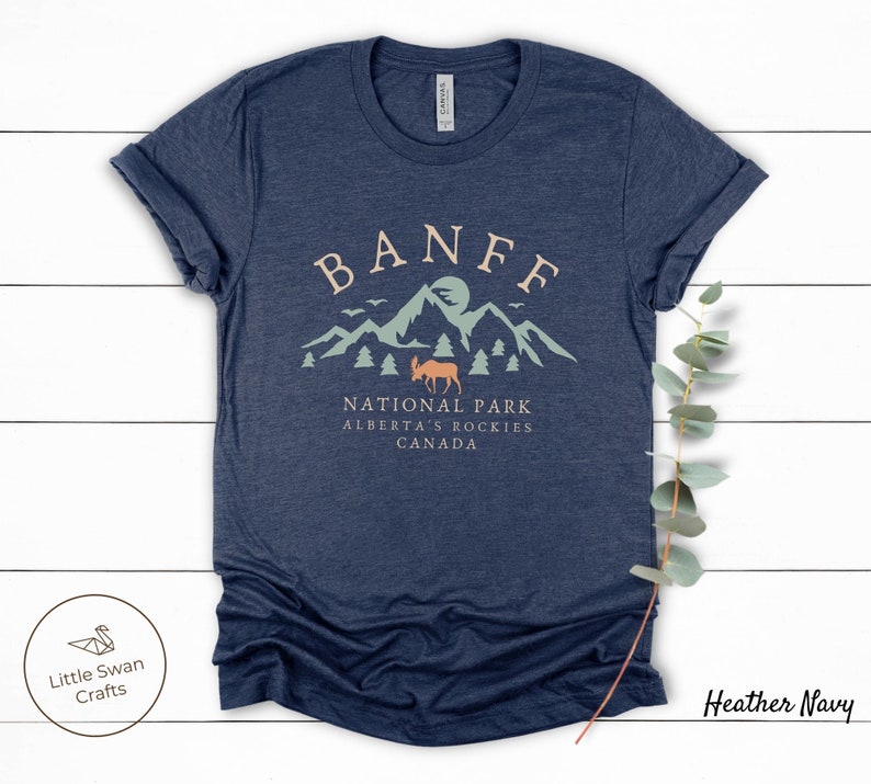 May include: Heather navy blue t-shirt with a graphic design of mountains, a moose, and the text "Banff National Park, Alberta's Rockies, Canada".