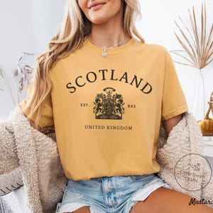 Pode incluir: Camiseta mostarda com a palavra "SCOTLAND" em preto, arqueada na parte superior. Abaixo, um bras&atilde;o com "EST. 843" e "UNITED KINGDOM". A camiseta &eacute; combinada com shorts jeans. A camiseta &eacute; de cor mostarda.
