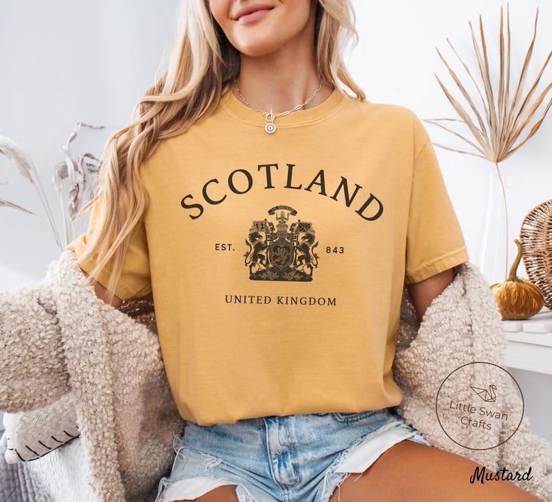 Pode incluir: Camiseta mostarda com a palavra "SCOTLAND" em preto, arqueada na parte superior. Abaixo, um bras&atilde;o com "EST. 843" e "UNITED KINGDOM". A camiseta &eacute; combinada com shorts jeans. A camiseta &eacute; de cor mostarda.