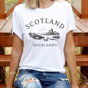 May include: White t-shirt featuring a black illustration of a Scottish Highlands scene, with a castle, sheep, and the words "SCOTLAND HIGHLANDS". The top has short sleeves and is made of a soft fabric.