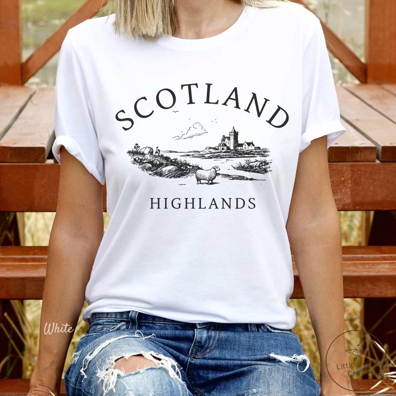 May include: White t-shirt featuring a black illustration of a Scottish Highlands scene, with a castle, sheep, and the words "SCOTLAND HIGHLANDS". The top has short sleeves and is made of a soft fabric.