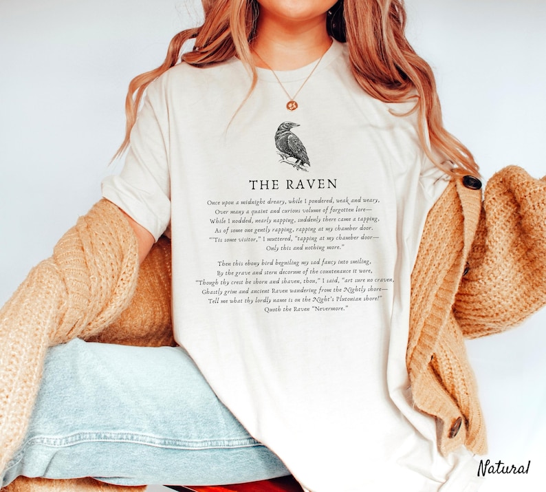 Puede incluir: Una camiseta blanca con una ilustraci&oacute;n de cuervo negro y el texto "THE RAVEN" impreso en la parte delantera. El texto debajo de la ilustraci&oacute;n del cuervo es un extracto del poema "El Cuervo" de Edgar Allan Poe.