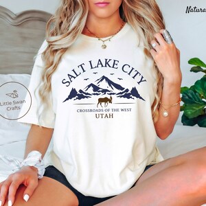 May include: White t-shirt with a blue graphic design that says "Salt Lake City" with a mountain range and a moose. The text below the graphic says "Crossroads of the West Utah".