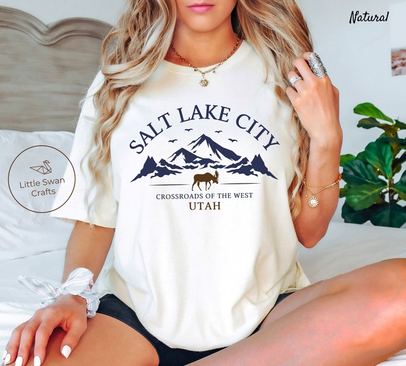 May include: White t-shirt with a blue graphic design that says "Salt Lake City" with a mountain range and a moose. The text below the graphic says "Crossroads of the West Utah".