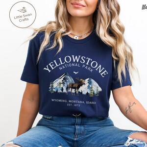 May include: Navy blue t-shirt featuring a Yellowstone National Park design. The graphic includes mountains, a moose, and the text "YELLOWSTONE NATIONAL PARK" with "WYOMING, MONTANA, IDAHO EST. 1872" below. The shirt is a short-sleeved crew neck.