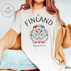May include: A white t-shirt with a graphic design featuring a circle of flowers, foxes, and moose with the text "FINLAND" and "suomi" in black.