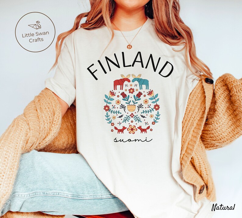 May include: A white t-shirt with a graphic design featuring a circle of flowers, foxes, and moose with the text "FINLAND" and "suomi" in black.