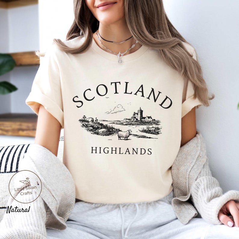 May include: A cream-coloured t-shirt with a black and white illustration of a Scottish landscape, including a castle, sheep, and the words "SCOTLAND HIGHLANDS". The shirt is made of a soft, natural fabric.