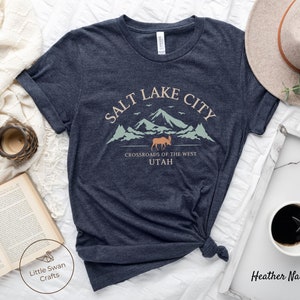 May include: Heather navy blue t-shirt with a graphic design of a mountain range with a moose and the text "Salt Lake City, Crossroads of the West, Utah".