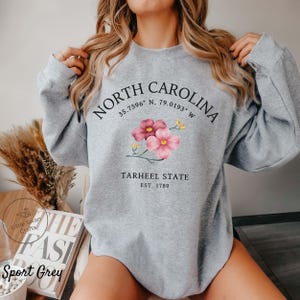 May include: Sport Grey sweatshirt with the text "NORTH CAROLINA" and coordinates, plus "TARHEEL STATE EST. 1789". The design includes pink flowers. The sweatshirt is a light grey color.