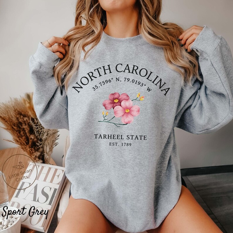 May include: Sport Grey sweatshirt with the text "NORTH CAROLINA" and coordinates, plus "TARHEEL STATE EST. 1789". The design includes pink flowers. The sweatshirt is a light grey color.