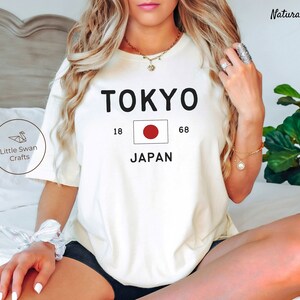 K&ouml;nnte beinhalten: Wei&szlig;es T-Shirt mit schwarzem Text, der "TOKYO" und "JAPAN" mit der japanischen Flagge dazwischen sagt. Die Zahlen 18 und 68 befinden sich auf beiden Seiten der Flagge.
