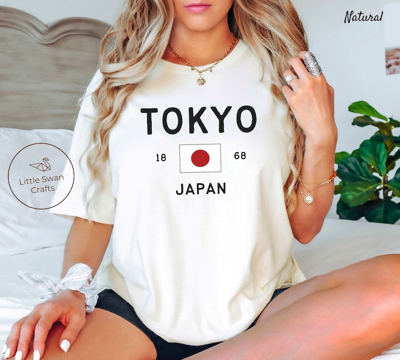 K&ouml;nnte beinhalten: Wei&szlig;es T-Shirt mit schwarzem Text, der "TOKYO" und "JAPAN" mit der japanischen Flagge dazwischen sagt. Die Zahlen 18 und 68 befinden sich auf beiden Seiten der Flagge.