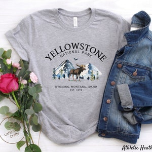 May include: A heather gray t-shirt featuring a Yellowstone National Park design. The graphic includes a moose, mountains, and trees. The text reads "Yellowstone National Park, Wyoming, Montana, Idaho, Est. 1872." A denim jacket is draped over the shirt.