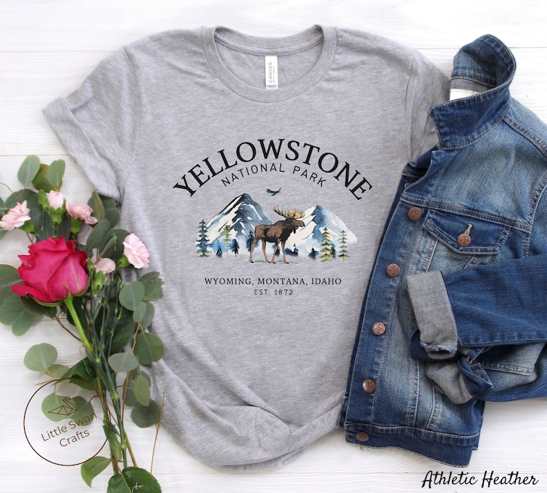 May include: A heather gray t-shirt featuring a Yellowstone National Park design. The graphic includes a moose, mountains, and trees. The text reads "Yellowstone National Park, Wyoming, Montana, Idaho, Est. 1872." A denim jacket is draped over the shirt.