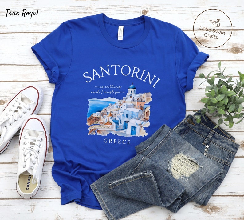 May include: Blue t-shirt with a white graphic of Santorini, Greece. The graphic includes a watercolor illustration of the town and the text "Santorini is calling and I must go".