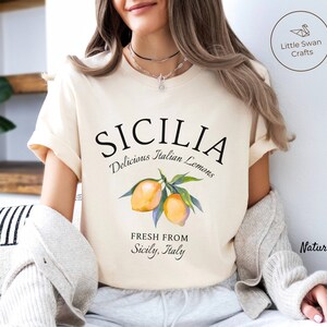 Peut inclure: Un t-shirt cr&egrave;me avec un texte noir qui dit "SICILIA Delicious Italian Lemons Fresh From Sicily, Italy" et une illustration aquarelle de citrons sur une branche.