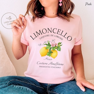 May include: Pink t-shirt with a graphic design of lemons and a lemon tree branch. The text on the shirt reads "Limoncello Liquore de Limone Fresco Costiera Amalfitana Prodotto Italiano".