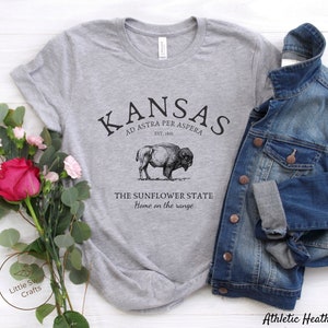 May include: Gray t-shirt with a black graphic design that says "Kansas" with the text "Ad Astra Per Aspera" and "Est. 1861" underneath. The graphic also includes a bison and the text "The Sunflower State" and "Home on the range".
