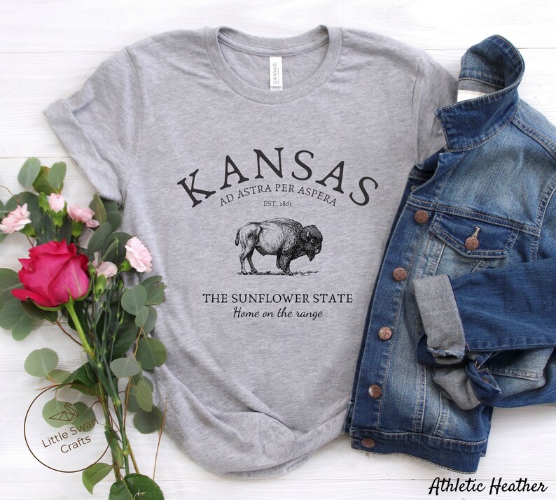 May include: Gray t-shirt with a black graphic design that says "Kansas" with the text "Ad Astra Per Aspera" and "Est. 1861" underneath. The graphic also includes a bison and the text "The Sunflower State" and "Home on the range".