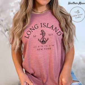 Op de afbeelding: Een heidekleurig T-shirt met een zwart grafisch ontwerp van een anker en de tekst "LONG ISLAND Est 1664 40.8&deg;N 73.3&deg;W NEW YORK"