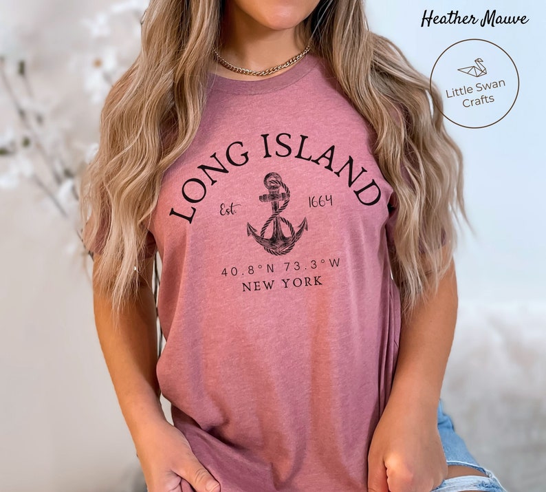 Op de afbeelding: Een heidekleurig T-shirt met een zwart grafisch ontwerp van een anker en de tekst "LONG ISLAND Est 1664 40.8&deg;N 73.3&deg;W NEW YORK"