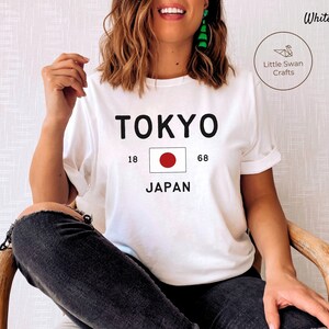 K&ouml;nnte beinhalten: Wei&szlig;es T-Shirt mit schwarzem Text, der "TOKYO" und "JAPAN" mit der japanischen Flagge dazwischen sagt. Die Zahlen 18 und 68 befinden sich auf beiden Seiten der Flagge.