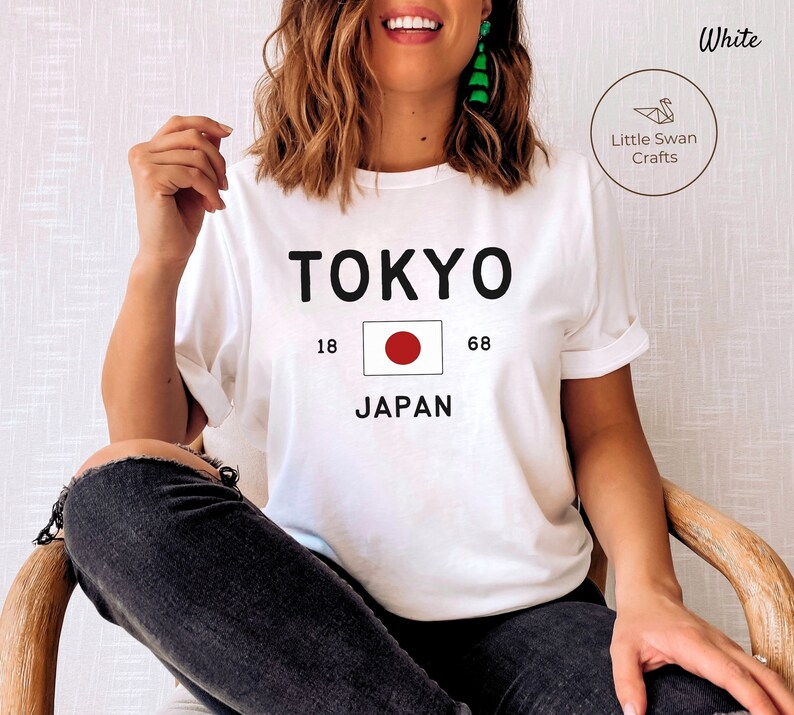 K&ouml;nnte beinhalten: Wei&szlig;es T-Shirt mit schwarzem Text, der "TOKYO" und "JAPAN" mit der japanischen Flagge dazwischen sagt. Die Zahlen 18 und 68 befinden sich auf beiden Seiten der Flagge.