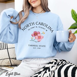 May include: Light blue sweatshirt with the text "NORTH CAROLINA" and coordinates, a floral design, and "TARHEEL STATE EST. 1789". The bottom of the sweatshirt has the text "Light Blue".
