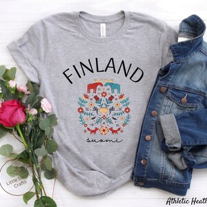 May include: A heather grey t-shirt with a black and white graphic design of a moose, flowers, and foxes. The text "FINLAND" is above the graphic and the text "suomi" is below the graphic.